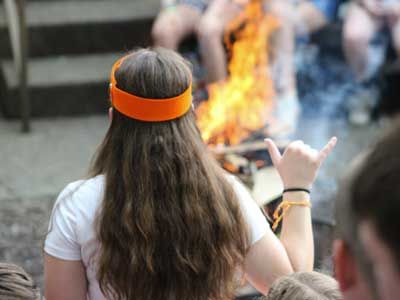 Young girl from behind, sitting in front of a fire wearing an orange headband.