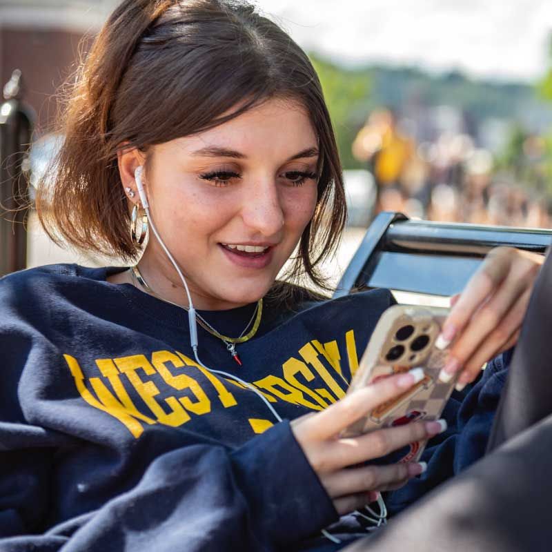 A young female WVU student sitting on a bench with her phone on the downtown campus.
